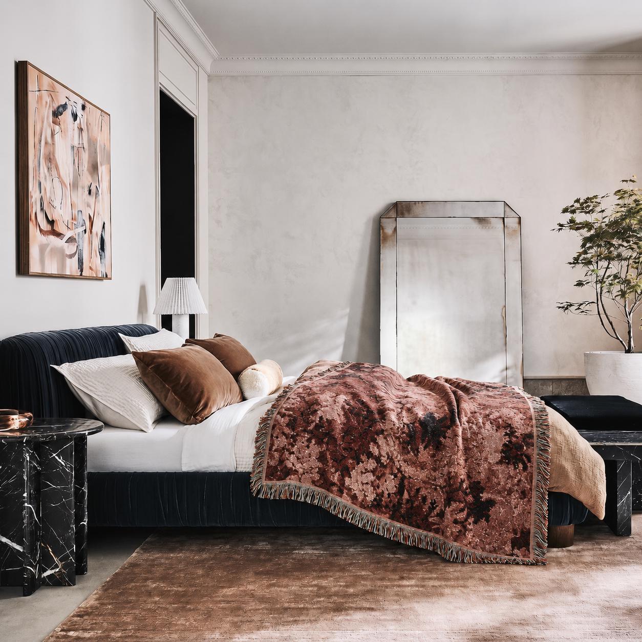 May include: A dark navy upholstered bed frame with white pillows, brown velvet cushions, and a textured, fringed throw blanket in shades of rust and brown. A black marble side table stands beside the bed, which is set in a room with a large mirror.